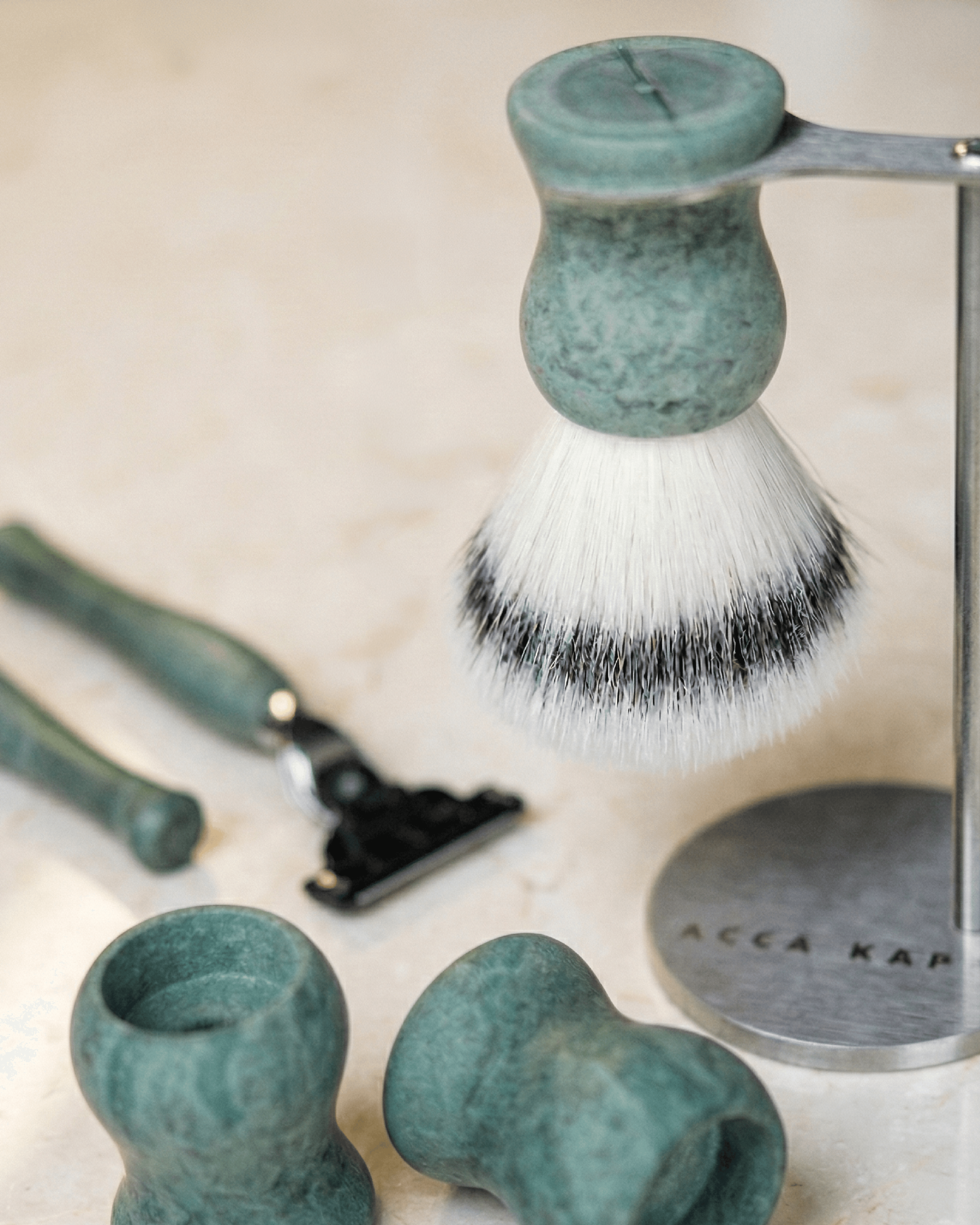 A close-up of a shaving set with a sculpted sage green and white shaving brush on a metal stand, alongside matching sage green accessories on a light beige marble surface. The metal base of the stand is engraved with the brand name 'ACCA KAR'.